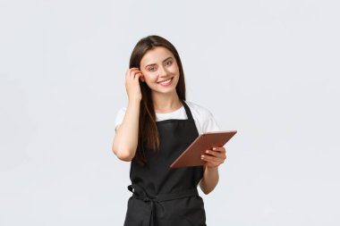 Market çalışanları, küçük işletmeler ve kahve dükkanları konsepti. Siyah önlüklü sevimli bayan barista kameraya gülümsüyor, kafeterya siparişlerini yönetmek için dijital tablet kullanıyor.