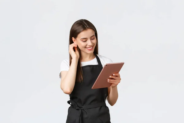 Empregados de mercearia, conceito de pequenas empresas e cafés. Amigável bonito barista feminino em avental preto sorrindo para a câmera, usando tablet digital para gerenciar encomendas de café — Fotografia de Stock