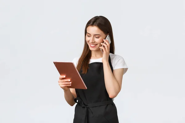 Empregados de mercearia, conceito de pequenas empresas e cafés. Bonito sorrindo vendedora em avental preto confirmar ordem, falando com o cliente no telefone e olhando para tablet digital — Fotografia de Stock