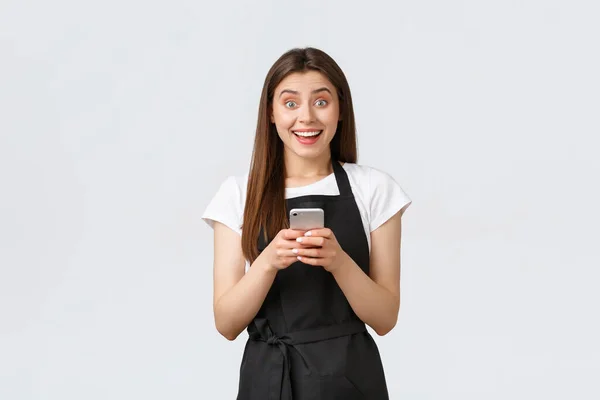 Grocery store employees, small business and coffee shops concept. Enthusiastic smiling cute female barista receive good news, rejoicing as holding mobile phone, stand white background