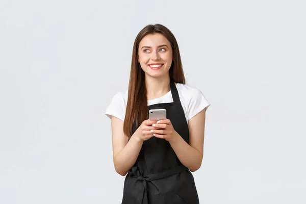 Grocery store employees, small business and coffee shops concept. Smiling attractive waitress in black apron laughing and looking left while holding mobile phone, white background — Stock Photo, Image