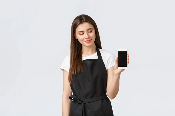 Zaměstnanci obchodu s potravinami, koncept malých obchodů a kaváren. Krásná mladá žena kavárna pracovník v černé zástěře inzerovat mobilní aplikace, ukazující displej smartphone, stojí bílé pozadí — Stock fotografie