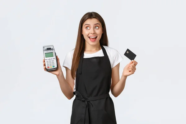 Grocery store employees, small business and coffee shops concept. Enthusiastic cheerful barista, saleswoman in black apron show POS terminal with credit card, looking left amused smile