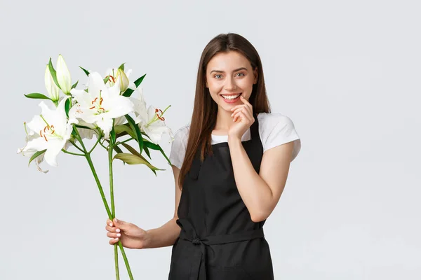 Einkaufen, Mitarbeiter und Kleinunternehmerkonzept. Lächelnde attraktive Verkäuferin im Blumenladen mit Schürze, sieht glücklich aus, als würde sie einen Strauß aus weißen Lilien machen, stehender weißer Hintergrund — Stockfoto
