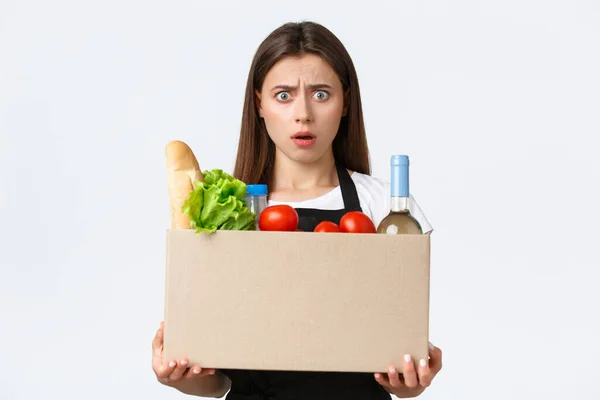 Empleados, entrega y pedidos en línea, concepto de supermercados. Cajero femenino conmocionado y confundido, vendedora sosteniendo paquete de bienes, caja con comestibles y frunciendo el ceño frustrado —  Fotos de Stock