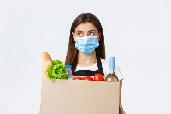 Covid-19 sociale afstand, levering en boodschappen doen tijdens het coronavirus concept. Jonge aangename verkoopster, kassier in medisch masker behandeling over boodschappen aan klant — Stockfoto