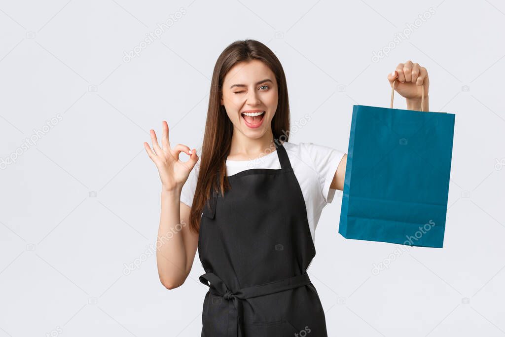 Grocery store employees, small business and coffee shops concept. Friendly smiling pretty cashier in black apron handing over paper bag with your gift purchase, show okay sign and wink cheeky