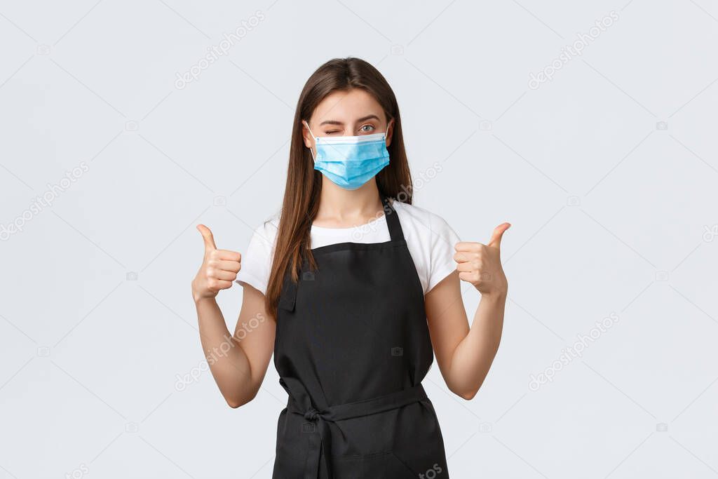 Covid-19 social distancing, cafe employees, coffee shops and coronavirus concept. Confident pretty barista in medical mask wink and show thumbs-up, assure all preventing virus measures are followed