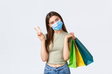 Sosyal uzaklık, koronavirüs ve alışveriş, moda konsepti. Cavid-19 sırasında tıbbi maske takan aptal, şık bir kadın alışveriş merkezlerinden alışveriş yapıyor. Barış işareti ve satın alınmış eşyalarla dolu çantalar gösteriyor.