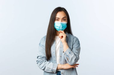 Sosyal mesafeli yaşam tarzı, covid-19 salgını günlük yaşam ve eğlence kavramı. Tıbbi maske takan Asyalı kuşkulu kızın endişeleri veya tereddütleri vardır. Çenesine dokun ve kameraya iyi bak.