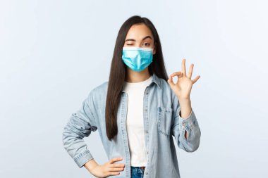 Sosyal mesafeli yaşam tarzı, covid-19 salgını günlük yaşam ve eğlence kavramı. Mutlu, sakin ve kendinden emin, tıbbi maskeli Asyalı kız, göz kırp ve onay işareti yap, kalite garantisi ver.