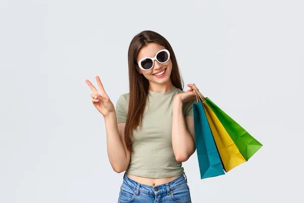 Shopping mall, lifestyle, tourism and fashion concept. Stylish young woman enjoying buying clothes in stores, tourist shop, holding bags and showing peace sign, white background — Stock Photo, Image