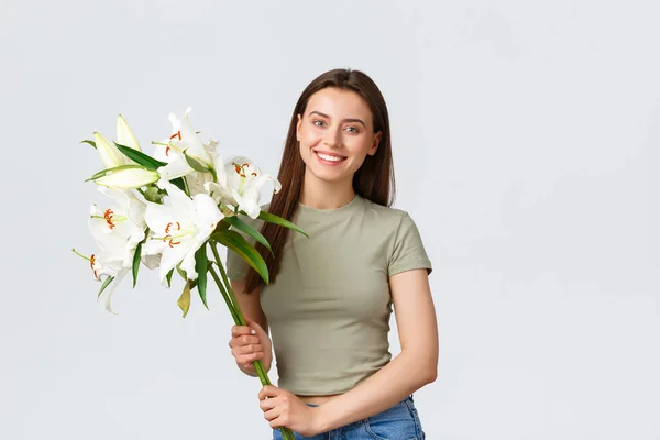 Happy good-looking young caucasian woman receive bouquet delivery, smiling at camera as holding flowers. Girl buy lilies for her home, standing white background pleased