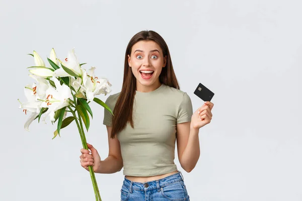 幸せな柔らかい若い女性は、クレジットカードで花の配達の花束を注文しました,笑顔と美しい白いユリを保持.女の子はインターネットでの購入のために支払い、白い背景に立って — ストック写真
