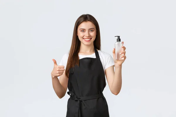 Grocery store employees, small business and coffee shops concept. Friendly pleasant female cafe worker, barista or saleswoman in black apron show thumb-up and hand sanitizer