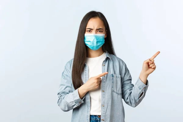 Social distanslivsstil, covid-19 pandemisk vardag och fritidskoncept. Missnöjd grimacing, arg asiatisk flicka i medicinsk mask rynkar pannan som pekar finger rätt — Stockfoto