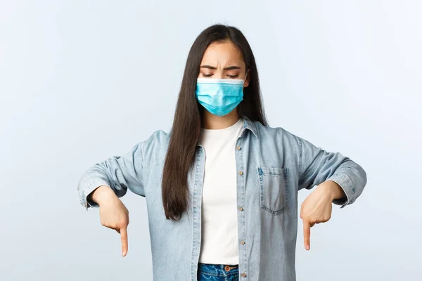 Social distanslivsstil, covid-19 pandemisk vardag och fritidskoncept. Allvarligt oroad asiatisk kvinna i medicinsk mask, tittar och pekar ner med missnöjd ansikte — Stockfoto