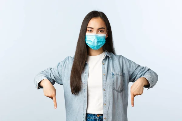 Estilo de vida social distanciamento, covid-19 pandemia vida cotidiana e conceito de lazer. Confiante e determinado jovem mulher asiática em máscara médica apontando os dedos para baixo em roupa casual — Fotografia de Stock