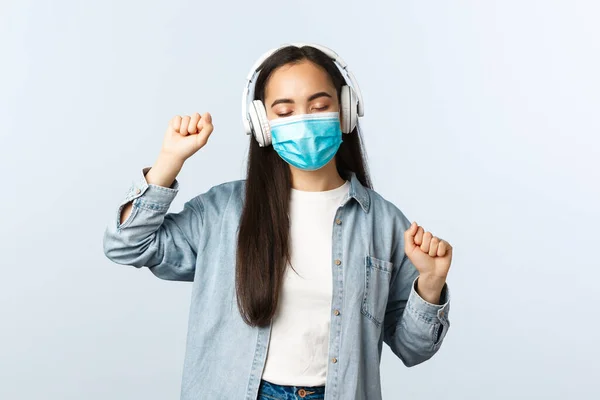 Social distancing lifestyle, covid-19 pandemic and self-isolation leisure concept. Happy upbeat asian girl in medical mask, listening music in wireless headphones and dancing — Stock Photo, Image