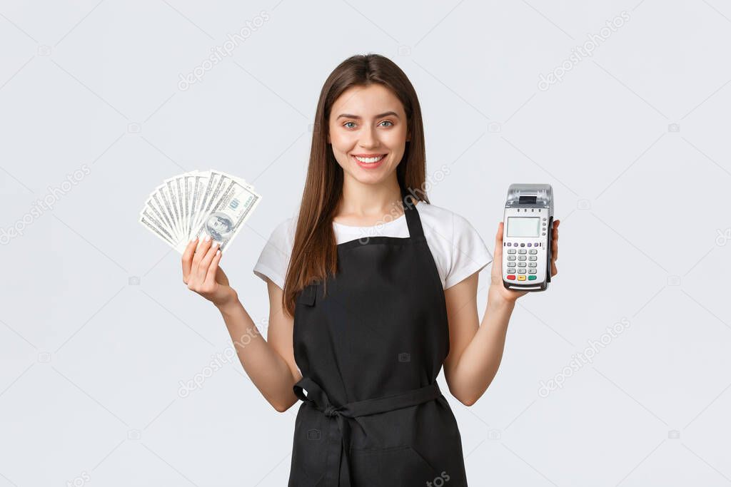 Grocery store employees, small business and coffee shops concept. Cheerful pretty female barista, cafe worker in black apron showing two ways of easy payment, POS terminal and cash