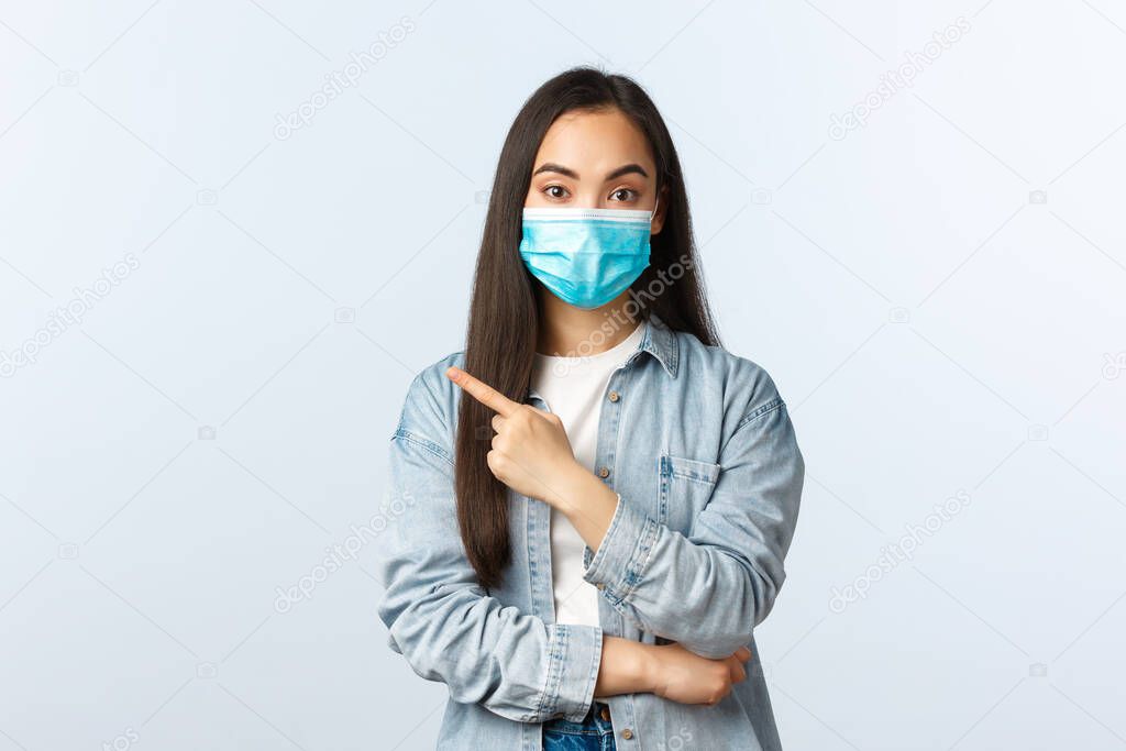 Social distancing lifestyle, covid-19 pandemic everyday life and leisure concept. Attractive korean girl in medical mask give advice, recommend visit store, pointing finger left at banner