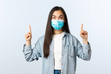 Social distancing lifestyle, covid-19 pandemic everyday life and leisure concept. Intrigued excited cute asian girl in medical mask pointing and looking up with enthusiastic happy face clipart