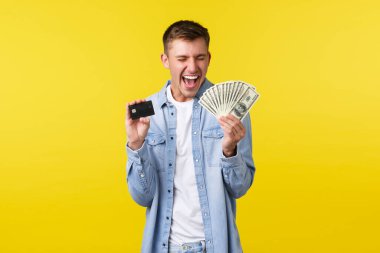 Heyecanlı yakışıklı sarışın adam para ve kredi kartı gösteriyor, evet diye bağırıyor, seviniyor, başarıyı kutluyor, ekstra para alıyor, piyango kazanıyor, sarı arka planda duruyor.