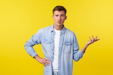 Yaşam tarzı, insan duyguları ve yaz tatili konsepti. Kafası karışmış ve dikkatsiz sarışın adam omuz silkiyor ve el kaldırıyor. Hayal kırıklığına uğramış, ne olduğunu anlayamıyor, sarı arka planda duruyor.