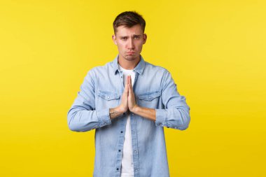 Yaşam tarzı, insan duyguları ve yaz tatili konsepti. Üzgün ve üzgün yakışıklı aptal adam somurtuyor, özür diliyor, iyilik diliyor, üzgün hissediyor, sarı arka planda duruyor.