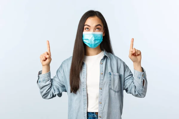 Social distancing lifestyle, covid-19 pandemic everyday life and leisure concept. Intrigued excited cute asian girl in medical mask pointing and looking up with enthusiastic happy face — Stock Photo, Image