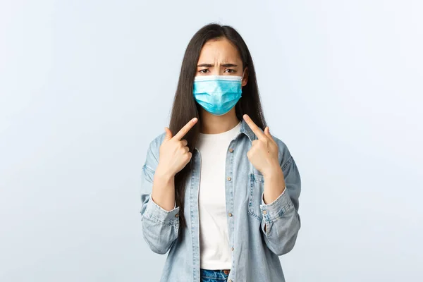 Social distanslivsstil, covid-19 pandemisk vardag och fritidskoncept. Missnöjd och störd asiatisk flicka pekar på ansiktet i medicinsk mask som inte gillar att bära den på sommaren — Stockfoto