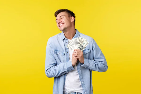 Concepto de inversión, compras y finanzas. Feliz hombre guapo rubio encantado, presionando el dinero en el corazón y sonriendo alegre, emocionado como la lotería ganadora, tiene pago de bonificación, fondo amarillo — Foto de Stock