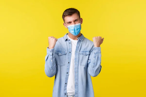 Concept of covid-19 pandemic outbreak, lifestyle during coronavirus social distancing. Happy handsome guy in medical mask, fist pump and saying yes, rejoicing over win, feeling lucky — Stock Photo, Image