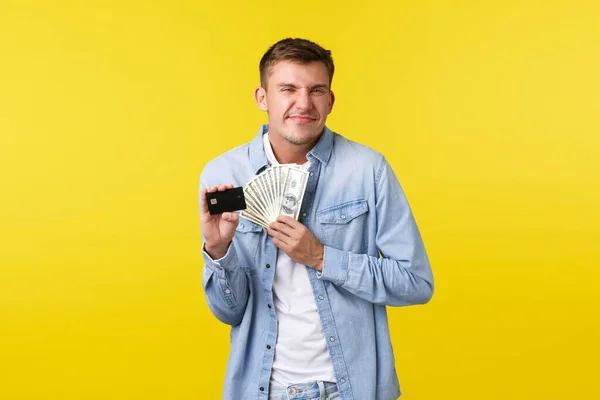 Alegre y encantado chico rubio feliz mirando emocionado, sonriendo satisfecho como recibir pago en efectivo extra, la celebración de la tarjeta de crédito con dinero, de pie fondo amarillo optimista —  Fotos de Stock