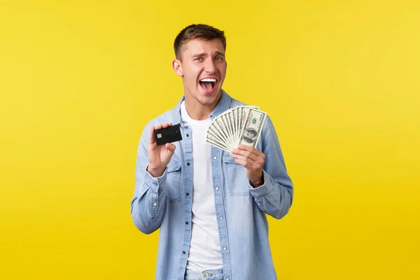 Pobavený šťastný blonďák radující se z placení, ukazující kreditní kartu a peníze. Muž triumfuje jako vítěznou loterii, obdrží první výplatu, získat peníze na letní dovolenou, žluté pozadí — Stock fotografie
