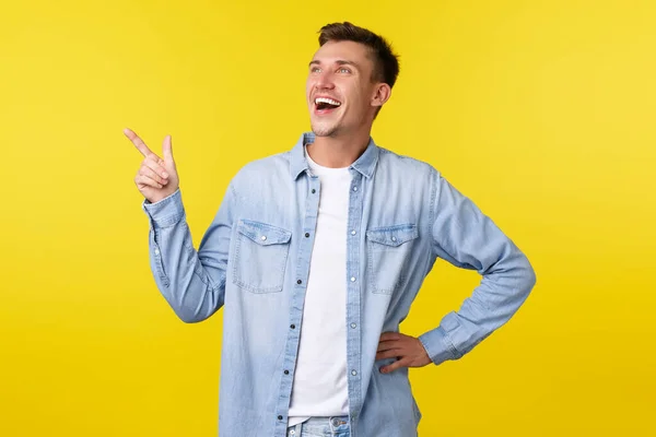 Gelukkige charismatische blonde man wijzen linksboven, lachen over grappige promo banner, genieten van geweldig evenement, plezier hebben en staan over gele achtergrond — Stockfoto