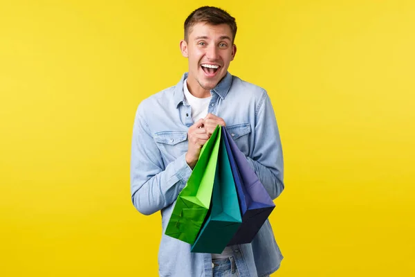 Concepto de compras, ocio y descuentos. Sonriente chico guapo feliz shopaholic, le gusta comprar cosas en ofertas especiales, la celebración de bolsas con expresión satisfecha, fondo amarillo — Foto de Stock