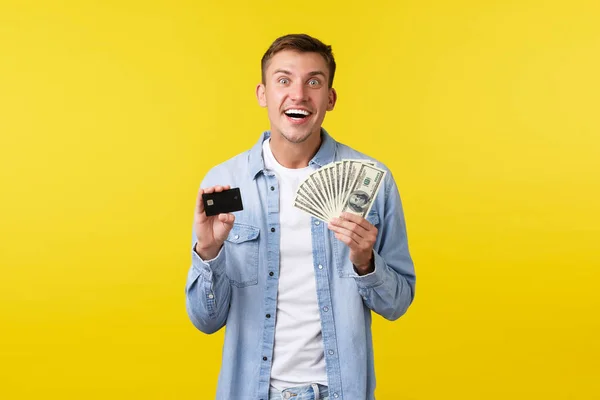 Emocionado hombre rubio sonriente feliz mirando emocionado y mostrando dinero con tarjeta de crédito, listo para pagar en efectivo por el producto, pagando por algo con expresión alegre, fondo amarillo — Foto de Stock