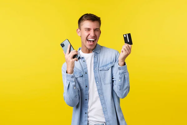 Technologie, style de vie et concept de publicité. Joyeux joyeux bel homme dansant comme fait un achat impressionnant en ligne, montrant la carte de crédit et le téléphone mobile comme se réjouissant, fond jaune — Photo