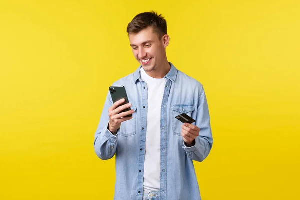 Teknoloji, yaşam tarzı ve reklam konsepti. Yakışıklı, güler yüzlü mutlu sarışın adam cep telefonu uygulamasına bakıyor, alışveriş sitesinde sipariş ya da teslimat için ödeme yapıyor, kredi kartı tutuyor, sarı arka plan — Stok fotoğraf