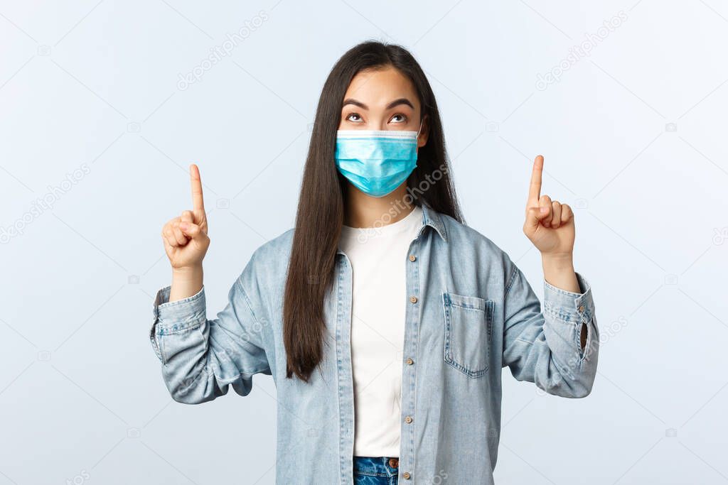 Social distancing lifestyle, covid-19 pandemic everyday life and leisure concept. Intrigued excited cute asian girl in medical mask pointing and looking up with enthusiastic happy face