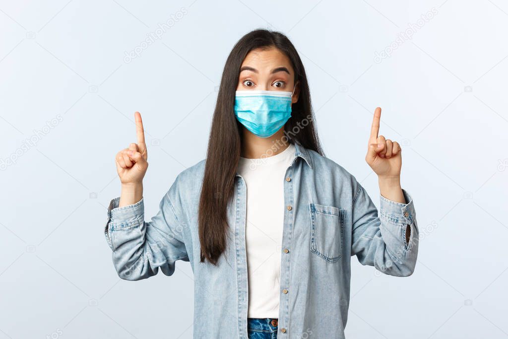 Social distancing lifestyle, covid-19 pandemic everyday life and leisure concept. Surprised and interested female student in medical mask hear about new interesting program, pointing fingers up
