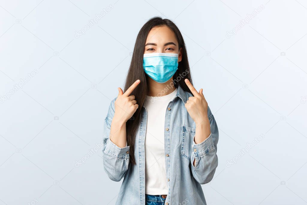 Social distancing lifestyle, covid-19 pandemic everyday life and leisure concept. Cheerful friendly-looking asian woman advice wear medical masks, pointing at herself as pefect example