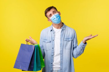 Coronavirus sırasında covid-19 salgını, alışveriş ve yaşam tarzı kavramı. Kararsız, kafası karışmış, tıbbi maskeli yakışıklı, habersiz omuz silkmiş ve dükkandan çanta taşıyor.