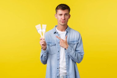 Hobiler, eğlence ve insanların yaşam tarzı konsepti. Şüpheci ve hayal kırıklığına uğramış seçici adam resme işaret ediyor kötü kalite fırçalar, sırıtıyor ve etkilenmemiş görünüyor, sarı arka planda şikayet ediyor.