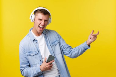Boş zaman, teknoloji ve uygulama reklam kavramı. Heyecanlı, mutlu, sarışın bir adam harika müzik dinliyor, kulaklıkla şarkılar dinliyor, cep telefonu tutuyor ve görünmez gitar çalıyor.