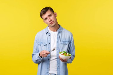 Sağlıklı yaşam tarzı, insanlar ve yemek konsepti. Kasvetli ve üzgün yakışıklı adam üzgün hissediyor salata yiyor, başını eğiyor diyete katılmıyor mükemmel vücut için formda kalmaya çalışıyor, sarı arka plan