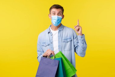 Coronavirus sırasında covid-19 salgını, alışveriş ve yaşam tarzı kavramı. Tıbbi maskeli genç düşünceli bir adam, parmağını kaldırıyor, öneri ya da fikri var, dükkandan çantalar alıyor.