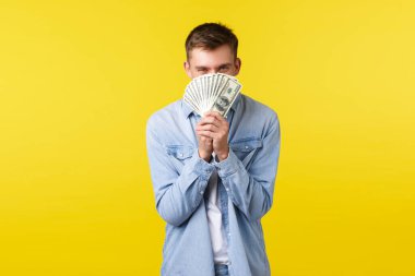 Yatırım, alışveriş ve finans konsepti. Neşeli genç adam parayı yüzüne karşı tutuyor, gülümseyen gözlerle bakıyor, zevk alıyor, piyangodan para kazanıyor, sarı arka planda.