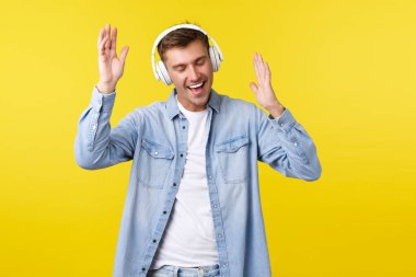 Yaşam tarzı, yaz tatili, teknoloji konsepti. Heyecanlı, sıradan kıyafetlerle yakışıklı adam, kablosuz kulaklıkla müzik dinliyor, dans ediyor ve gülümsüyor, favori şarkının keyfini çıkarıyor, sarı arka planda.